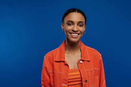 A young African American woman, in an orange shirt, beams happily at the camera against a blue...