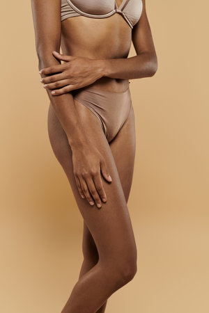 African American woman in tan bra and panties showcases self-care on a beige background.