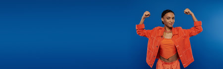 A young African American woman standing confidently in a vibrant orange shirt and pants against a blue background.の写真素材