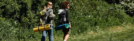 Two young women, one with long blonde hair and the other with short afro hair, hike through a wooded...