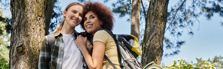 A young, multicultural lesbian couple smiles while hiking in the forest, surrounded by tall trees.の写真素材