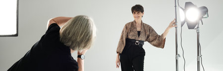 A middle-aged lesbian couple in a photo studio; one shoots with a camera, the other poses elegantly.