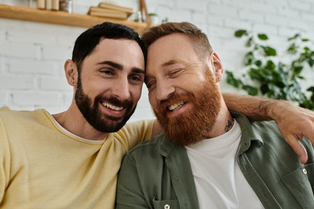 Two men, both with beards, relax on a couch and hugging at homeの写真素材