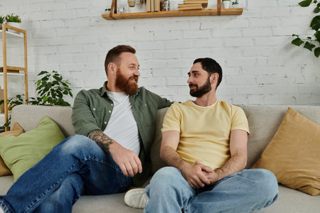 Bearded gay couple sitting on a couch, engaged in a heartfelt conversation in a cozy living room setting.の写真素材