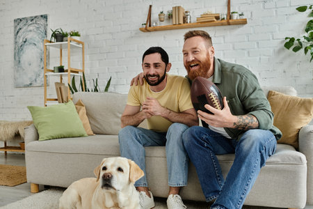 A man with a beard is seated on a couch next to a Labrador, relaxing and watching a sports match.の写真素材