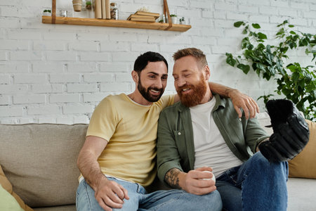 A pair of bearded men relax together on a couch, watching a sports match in their living room.の写真素材