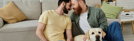 Two men, a bearded gay couple, sit on a couch, near a friendly Labrador dog in a cozy living room setting.の写真素材