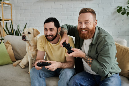 A bearded gay couple and their labrador dog sit on a couch intensively playing a video game together in the living room.の写真素材