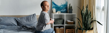 A man with inclusivity sits on the edge of his bed, holding a mug, with a window and a plant in the...
