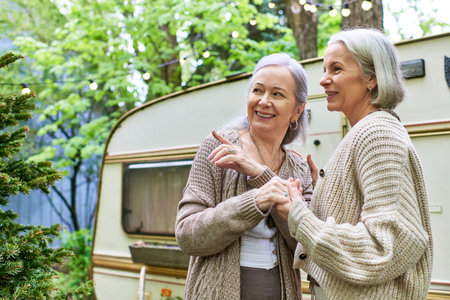 A lesbian couple smiles and holds hands in a verdant forest near a camping van.