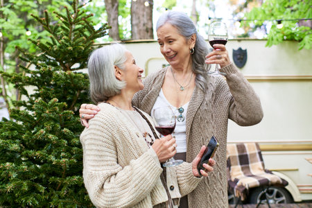 Two middle-aged women, a lesbian couple, stand outside their camping van, raising a toast to a fun trip in the woods.の写真素材