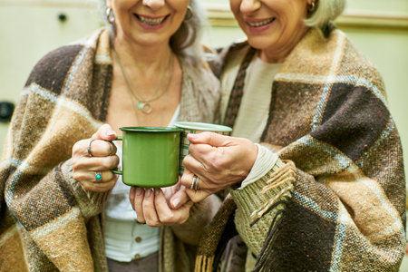 Two women wrapped in blankets, enjoying a warm drink on a camping trip.の写真素材