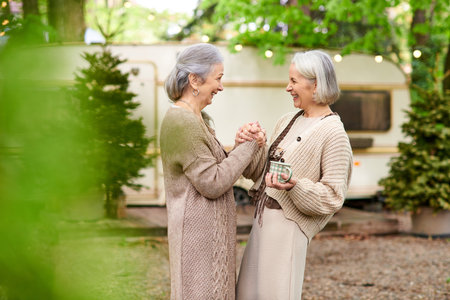 Two middle-aged women, a lesbian couple, stand near a camper van in a green forest setting, enjoying...