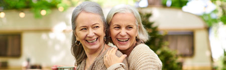 Two women in their 50s laugh and embrace while camping in a lush green forest.