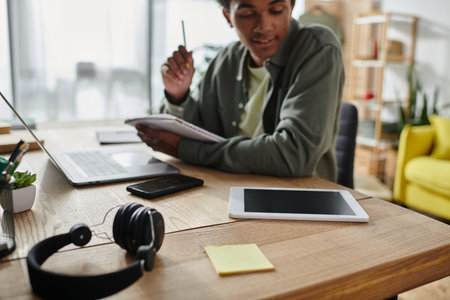 Young African American man immersed in online study with headphones and laptop at desk.