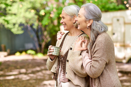 Two middle-aged women laugh together while enjoying a cup of coffee during a camping trip.