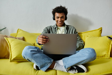 Young African American man engrossed in online study, seated on yellow couch with laptop.の写真素材