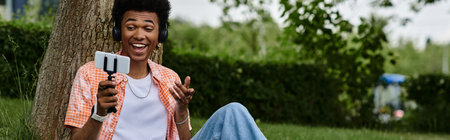 Young African American man sits under a tree, engrossed in his cell phone.の写真素材