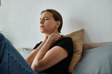Middle-aged woman sits on bed, hands on neck.