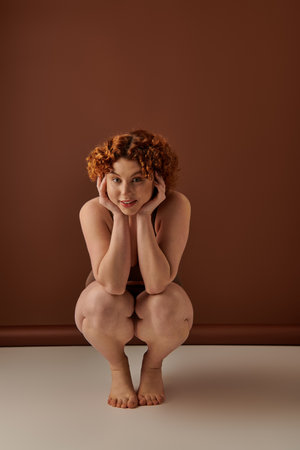 A young, curvy redhead woman crouches gracefully on the floor in her underwear against a brown backdrop.の写真素材