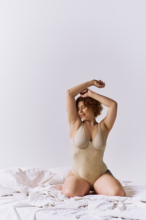 Young, curvy redhead woman in bodysuit sitting gracefully on bed against gray backdrop.の写真素材