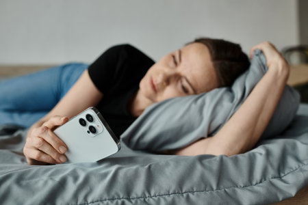 Middle-aged woman laying in bed, holding phone, lost in thought.の写真素材
