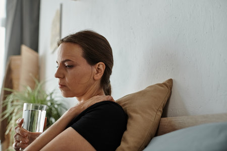 Middle-aged woman sitting on a bed holding a glass of water.の写真素材