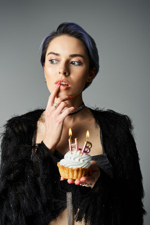 A young woman in stylish attire holds a cupcake with lit candles, a birthday girl ready to make a...