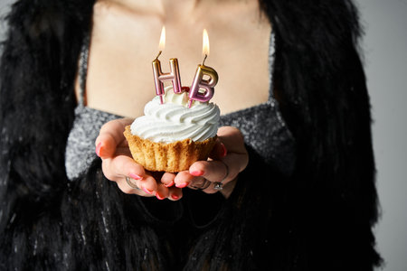 A stylish young woman with short dyed hair holding a cupcake with burning candles in a studio...