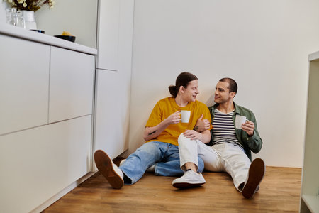 A young gay couple sits on the floor of their modern kitchen, enjoying a cup of coffee and each...