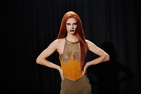 A drag queen in a shimmering gold corset and long red hair poses confidently in front of a black...