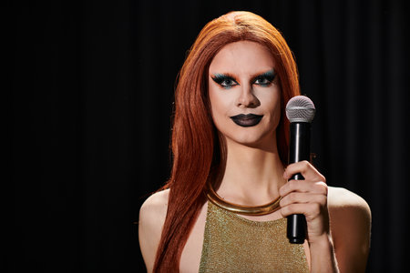 A drag queen with long red hair and bold makeup holds a microphone before a performance.の写真素材