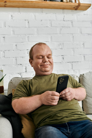 A man with inclusivity sits on a couch, casually dressed, and scrolls through his phone with a smile on his face.の写真素材