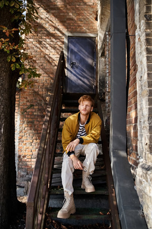 A young, red-haired man sits elegantly on a staircase in a bustling city.の写真素材