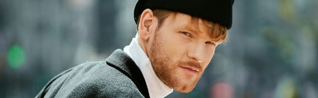 A young red-haired man with a beard, in debonair attire, strolling through the city wearing a black...