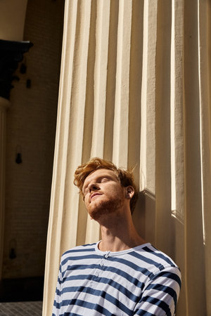 A stylish young man with red hair stands gracefully next to a richly colored curtain.の写真素材
