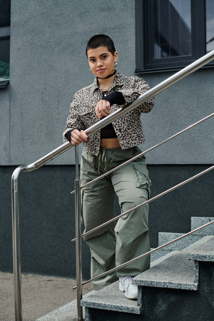 Stylish young woman with short hair balances on stair rail near modern building, showcasing daring...