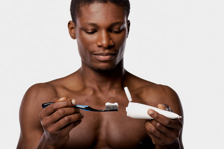 Shirtless African American man in studio with white background, holding a toothbrush and an...