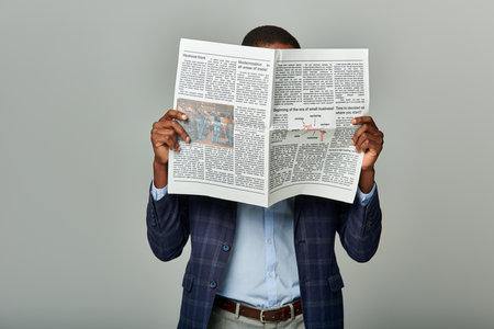 African American man in checkered blazer, reading newspaper attentively on gray background.の写真素材