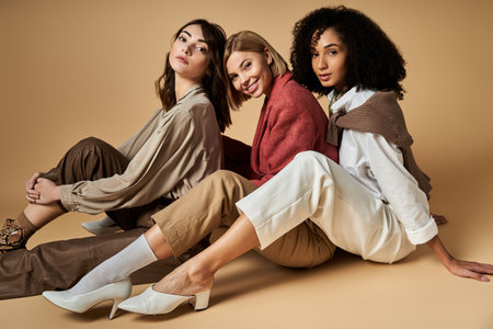 Three young, stylish women of different cultures sit on the ground, posing for a photo with a beige...