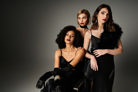 Three beautiful young women in black dresses posing gracefully for a stylish portrait on a gray backdrop.の写真素材