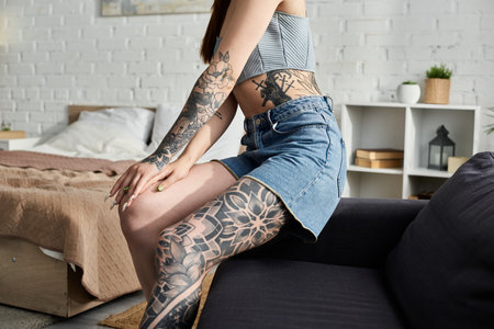 A young woman with tattoos sits on a couch in a modern apartment.の写真素材