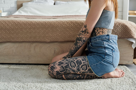 A young woman with intricate tattoos on her arms and legs sits on the floor of her modern apartment.の写真素材