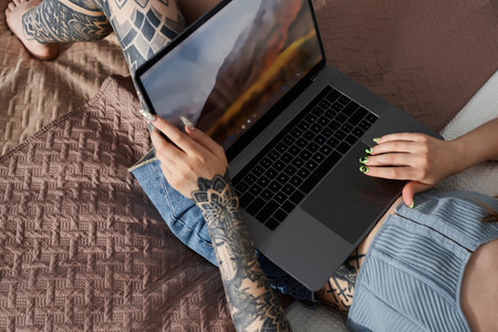 A young woman with tattoos sits on a couch in a modern apartment, browsing a laptop.