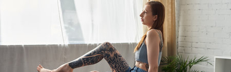 A young woman with tattoos sits by a window in her modern apartment, enjoying a cozy weekend.の写真素材