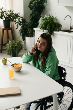 A young brunette woman with a disability sits in a wheelchair at her kitchen table, talking on her...