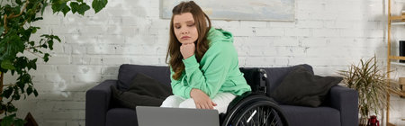 A young woman in a wheelchair sits on a couch in a living room, looking contemplative and staring at...
