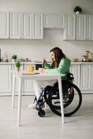 A young brunette woman with a disability sits in a wheelchair and eats breakfast at a white table in her home.の写真素材