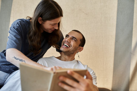 A young gay couple enjoys a cozy moment together, laughing while reading a book in their modern home.の写真素材