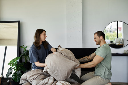 A young gay couple playfully fight with pillows on their bed in their bedroom.